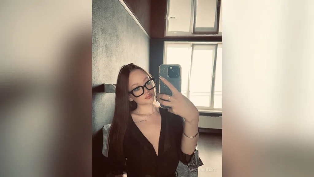 Young woman in a cream ribbed off-the-shoulder top taking a moody mirror selfie in a dimly lit room.