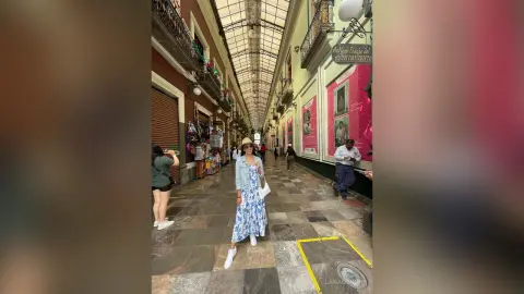 Stylish woman in a blue floral maxi dress walking through a historic sunlit European shopping arcade.