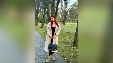 Red-haired woman in a beige trench coat and black dress posing on a park path during a sunny autumn day.