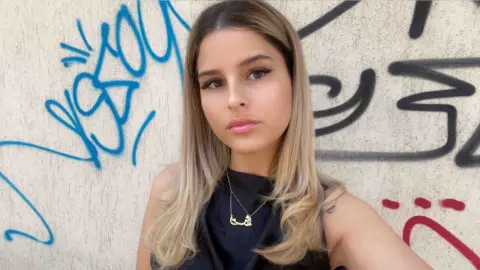 Young woman with blonde hair wearing a black sleeveless top posing against a blue and black graffiti wall.
