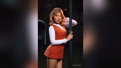 Stylish woman in a burnt orange pinafore dress posing playfully against a dark outdoor background.