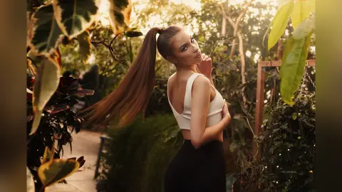 DelanaGrace poses outdoors with her long brown hair in a high ponytail, wearing a white crop top and black skirt.