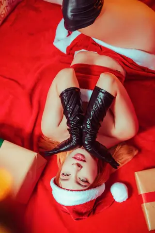 Young woman in a red and white Santa outfit and black gloves lying on red fabric surrounded by gifts.