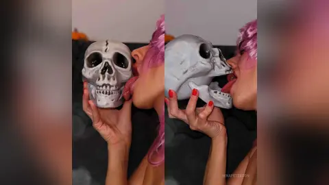 Woman with pink hair playfully licking a white skull prop in a moody, dark setting.