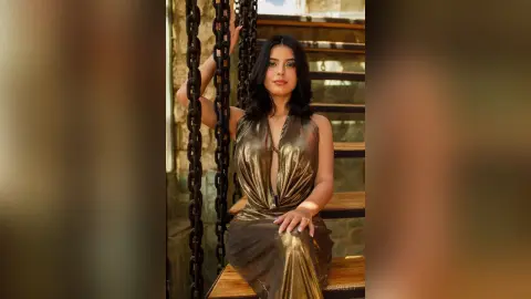 Elegant woman in a shimmering gold evening gown posing on wooden stairs with industrial chain decor.