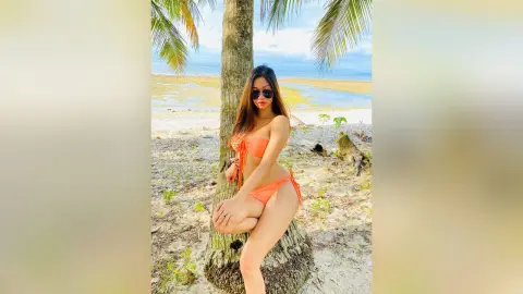 Stylish woman in an orange bikini posing by a palm tree on a sunny tropical beach.