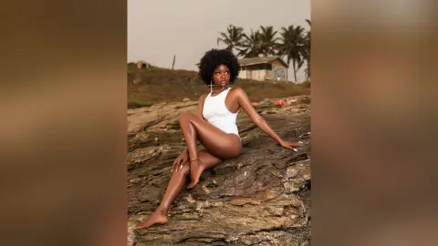 Young Black woman in a white bodysuit sitting on coastal rocks during a serene sunset.