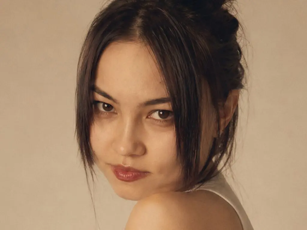 SeeBugl looks over her shoulder with dark hair framing her face against a plain tan background.