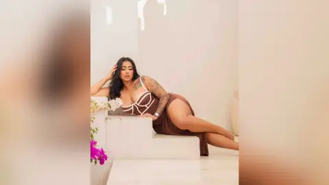 Elegant woman in a white lace bodysuit and brown wrap skirt lounging on white stairs in a bright, airy setting.