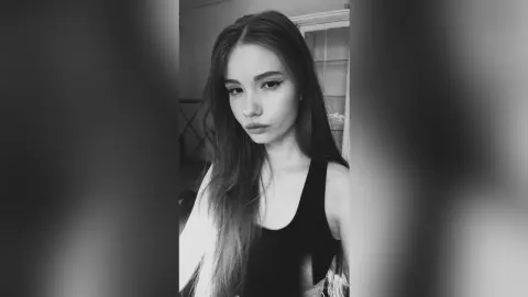 Moody black and white portrait of a young woman with long hair wearing a black tank top indoors.