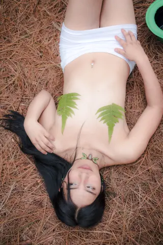 ScarletDaviis lies naked on a bed of pine needles, her long black hair spread out and her hands resting near her chest and waist.