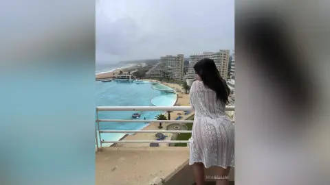 Woman in a white lace cover-up looking at a luxury resort swimming pool on a cloudy day.