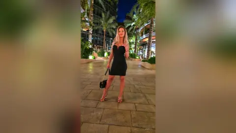 Elegant woman in a black cocktail dress and gold heels walking through a tropical resort at night.