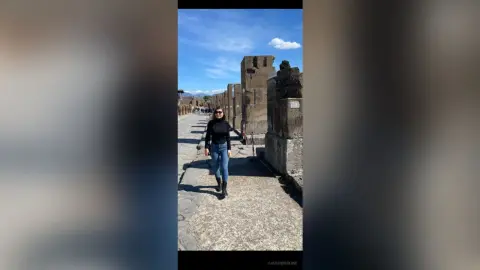 Woman in black sweater and jeans walking through ancient Roman ruins under a bright blue sky.