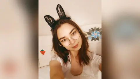 Young woman with bunny ears and glasses smiling in a cozy white bedroom setting.