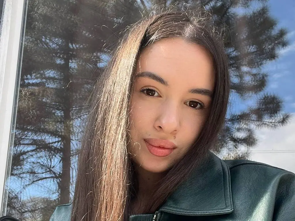 AmberPocasangre looks at the camera with long brown hair and a dark green leather jacket, with trees and a blue sky visible behind her.