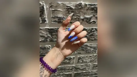 Close-up of a hand with blue and white marble patterned long almond nails against a textured stone wall.
