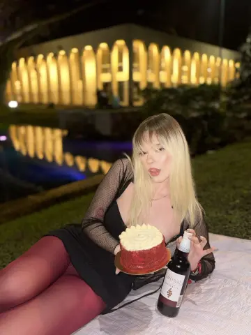 Blonde woman in a black dress holding champagne and cake during a romantic night picnic by a lit building.
