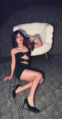 Young woman in a black cutout mini dress sitting on a white chair against a dark textured background.