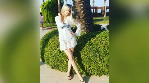 Blonde woman in a white lace summer dress standing in a sunny garden near palm trees.