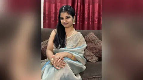 Sejuti sits on a brown couch wearing a light blue and silver shimmering saree, with long black hair and gold jewelry.