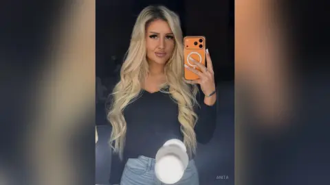 Anita poses for a mirror selfie wearing a black long-sleeved top and light blue denim jeans, letting her long blonde hair hang down.