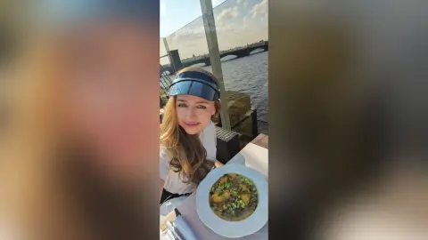 Smiling woman in a white top and sun visor enjoying a gourmet meal at a waterfront outdoor restaurant.