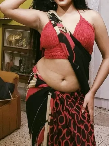 Woman in a red crop top and black patterned saree posing in a home setting.