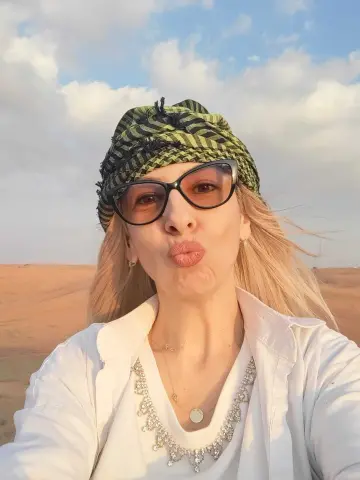 Blonde woman in white shirt and green patterned headscarf making a kissy face in a sunny desert landscape.