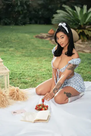 Young woman in blue gingham sundress sitting on a picnic blanket with strawberries in a lush garden setting.