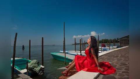 MissyJolie sits on a stone pier wearing a red dress, looking out over the water under a clear blue sky.