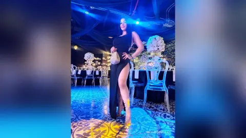 MeganneAllen poses in a black dress with a high thigh slit, standing in a decorated room with blue lighting and clear chairs.
