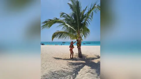 Woman in orange bikini standing on a sunny tropical beach next to a palm tree under a clear blue sky.