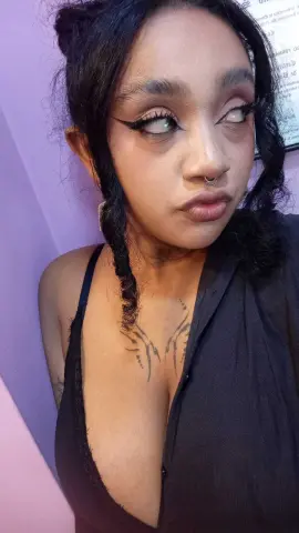 Young woman with winged eyeliner and dark braids wearing a black top in soft pink lighting.