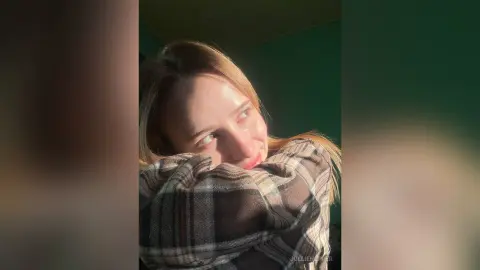 Smiling young woman in a cozy plaid shirt relaxing in warm, sunlit indoor lighting.