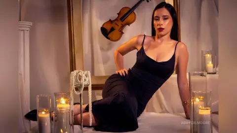 Elegant woman in a black evening gown sitting by candlelight near a violin in a moody, sophisticated setting.