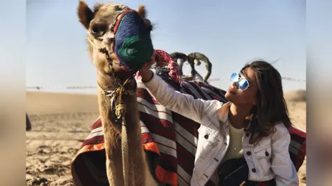 AnaMathers smiles while wearing blue sunglasses and a white jacket, reaching out to touch a camel's head in a desert setting.
