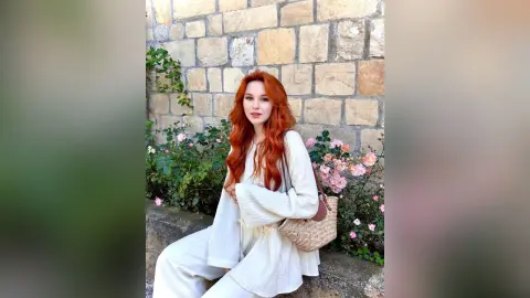 VetaFerr is a redhead woman wearing a white long-sleeved shirt and white pants, sitting outdoors by a stone wall and pink flowers.