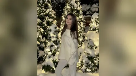 Elegant woman in a white sweater standing amidst snow-covered Christmas trees with glowing lights at night.