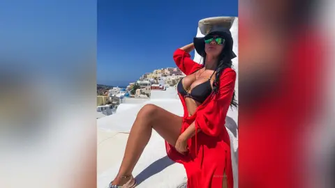 MissyJolie poses outdoors in a red wrap dress and black bikini top, wearing a black sun hat and green sunglasses.