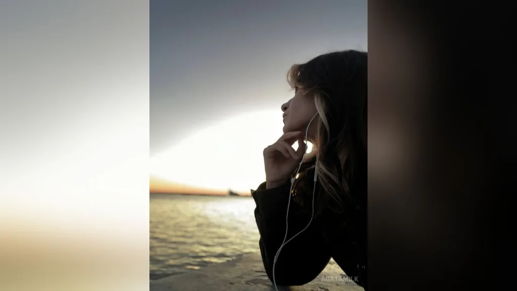 AgataMilk poses outdoors by the water at sunset wearing white earbuds.