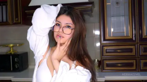 Young woman with glasses in a white blouse posing thoughtfully in a cozy kitchen setting.