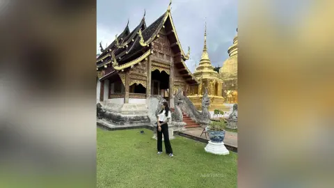 Woman in white top and black trousers standing before a traditional Thai temple under a cloudy sky.