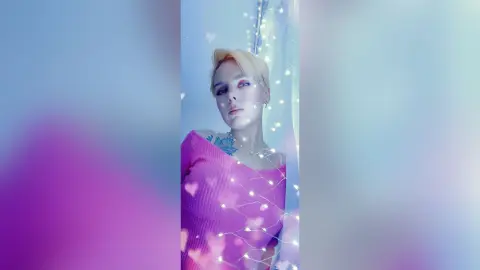 AvaLindt poses with short blonde hair, wearing a pink off-the-shoulder top and a blue chest tattoo, surrounded by soft white lights.