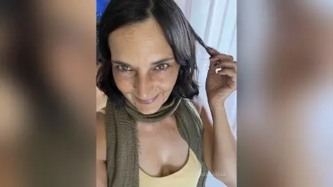 MichaelaBelle smiles while tucking a strand of her dark hair behind her ear, wearing a tan tank top and an olive green scarf.