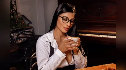 Young woman in white shirt and glasses enjoying a warm cup of coffee in a cozy cafe setting.