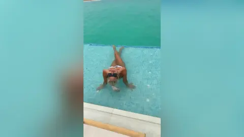 Relaxed blonde woman in a black bikini sunbathing on a turquoise swimming pool ledge during a summer vacation.