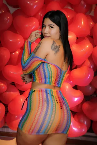 channeljonson — Young woman in colorful rainbow crochet outfit posing against a romantic red heart balloon background.