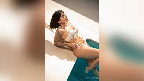 Tanned woman in a white bikini relaxing on a sunny poolside patio with tattoos.
