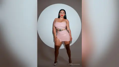 Confident woman in a pink strapless latex mini dress and brown thigh-high boots posing against a white circle.